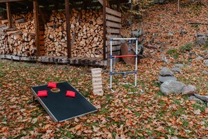 Un espacio al aire libre con una mesa negra, cojines rojos, una estructura de madera con leña apilada y un columpio metálico.