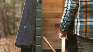 Une personne tenant un seau noir avec une anse en bois, se tenant à l’entrée d’un bâtiment en bois.
