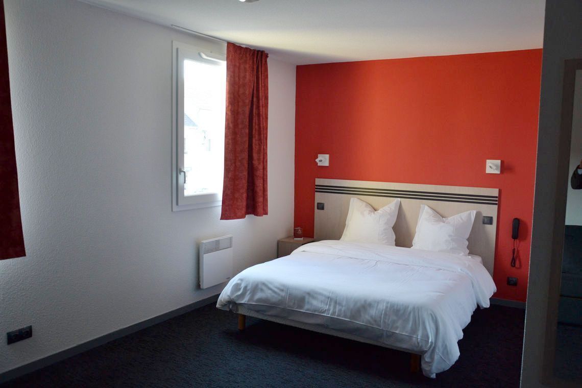 A hotel room with a bed, a window with red curtains, a radiator, and a red accent wall.
