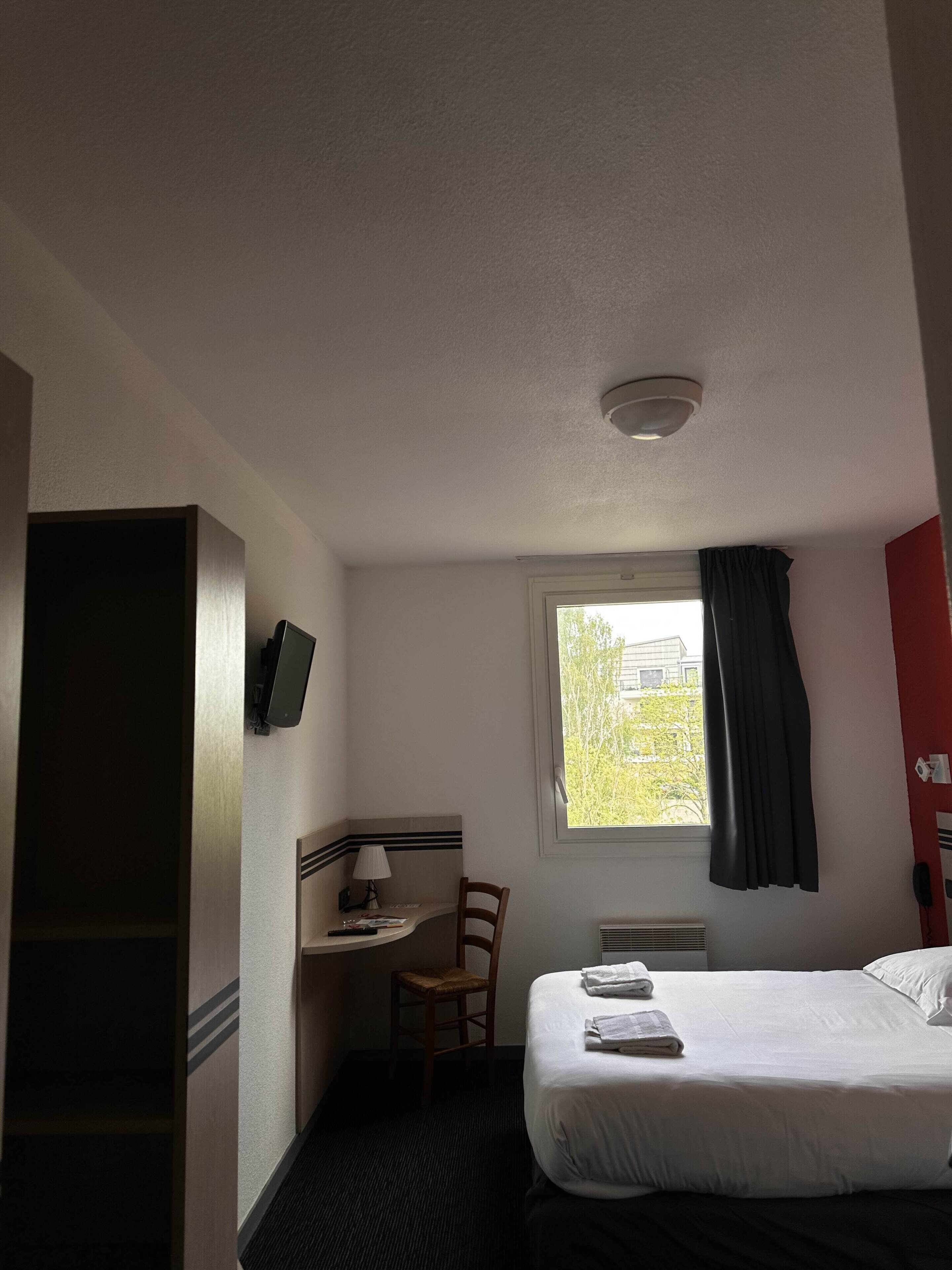 A hotel room with a bed, a desk with a chair, a window with curtains, and a television on the wall.