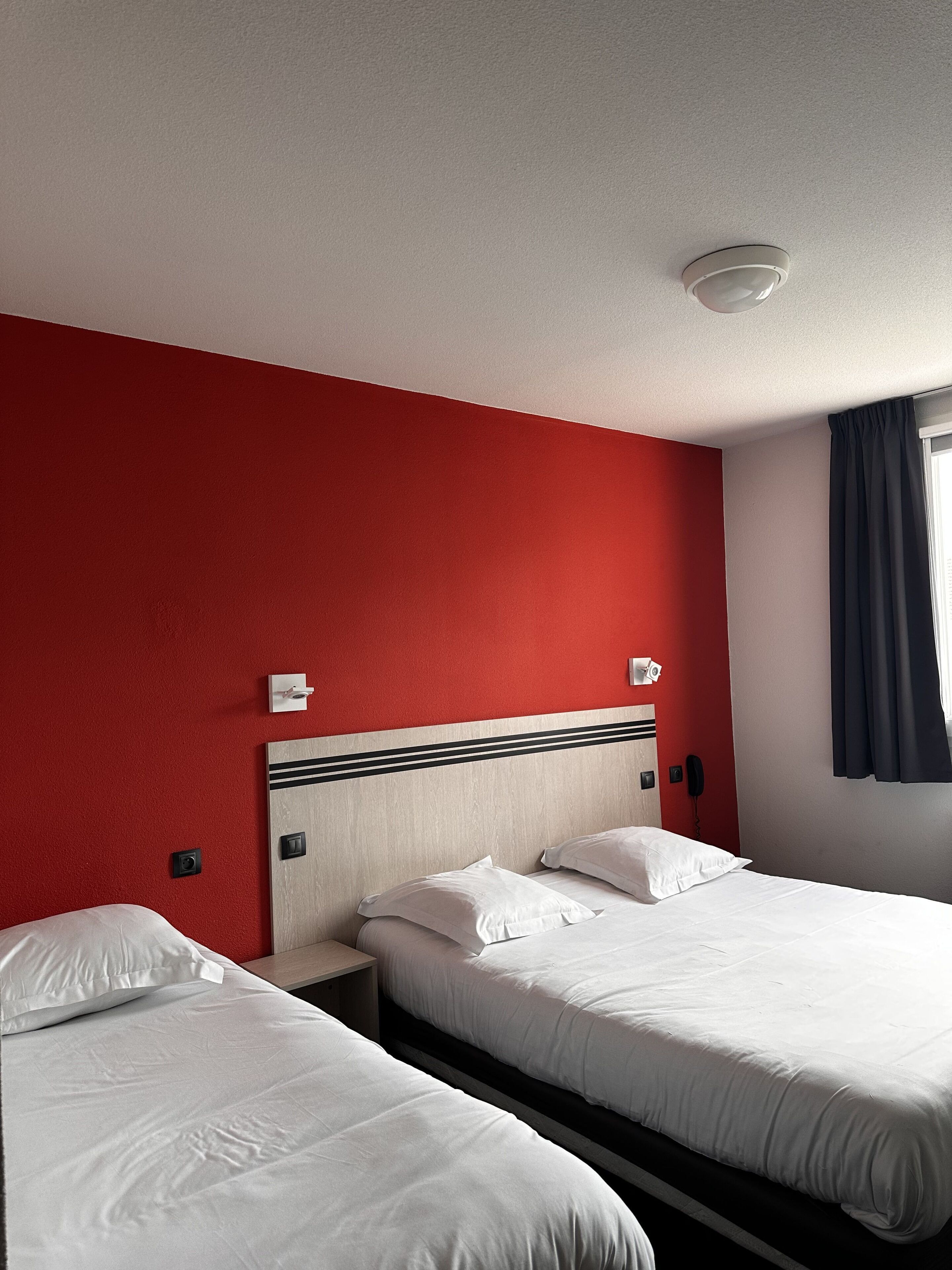 A hotel room with two single beds, a red accent wall, a wooden headboard, a white ceiling light, a telephone, and a window with dark curtains.
