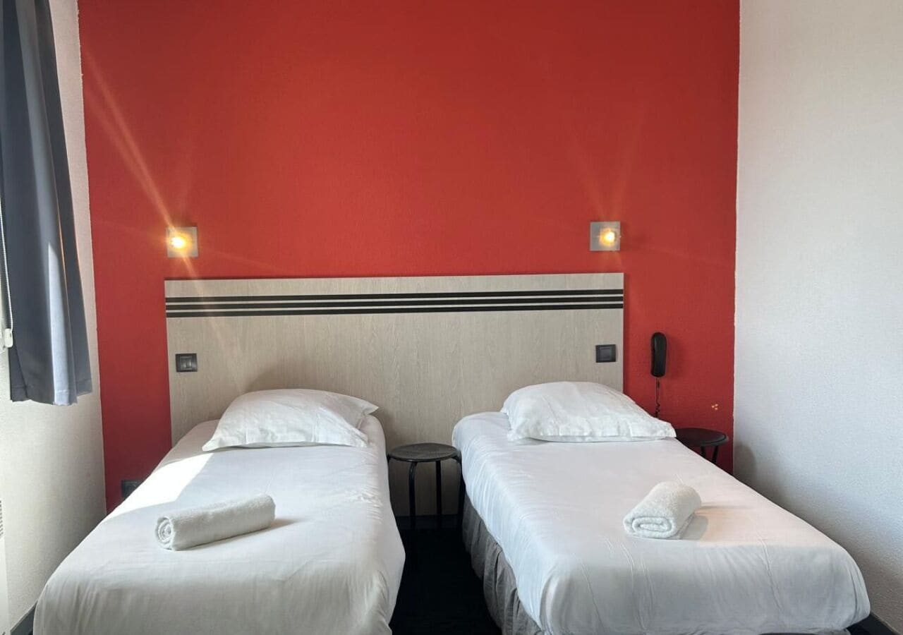 Une chambre d’hôtel avec deux lits simples, une tête de lit à motifs rayés, une petite table ronde et un téléphone fixé au mur.
