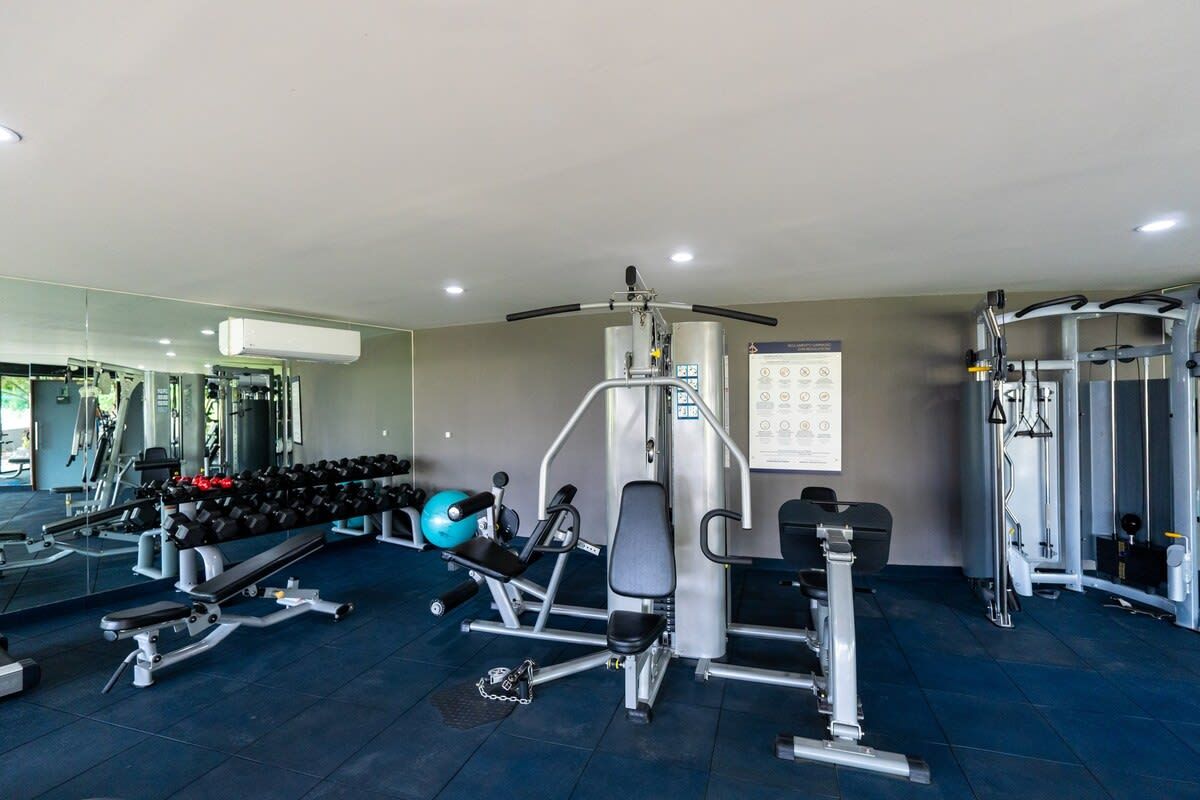 Un gimnasio con equipo de ejercicio variado, una prensa de banca, mancuernas y un espejo grande.