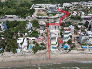 An aerial view of a coastal resort area with multiple buildings, including hotels and residential structures, alongside a beach with a swimming pool.