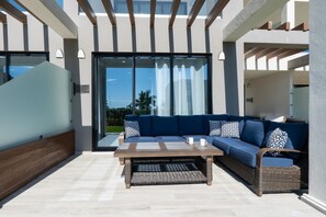 A modern outdoor seating area with a sofa and coffee table, large windows, and a view of the outdoors.