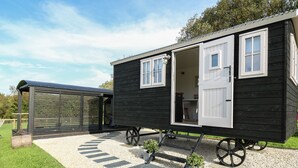 A black mobile home with a kitchen area visible through the open door, a small outdoor seating area, and a covered outdoor space. - Dragonfly Shepherd's Hut (Llanarth)