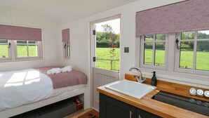 A bedroom with a bed, a window with a view of a green field, a small window with a curtain, and a kitchen with a sink and stove. - Dragonfly Shepherd's Hut (Llanarth)