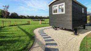A small, dark wooden house with a white window and a curved roof, situated on a gravel path in a grassy area. - Dragonfly Shepherd's Hut (Llanarth)