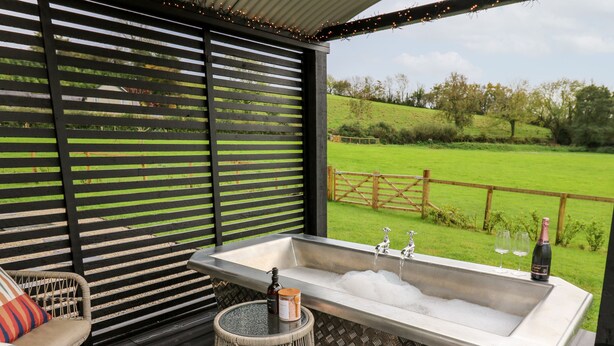 A covered outdoor area with a hot tub, wicker furniture, and a view of a green field. - Dragonfly Shepherd's Hut (Llanarth)