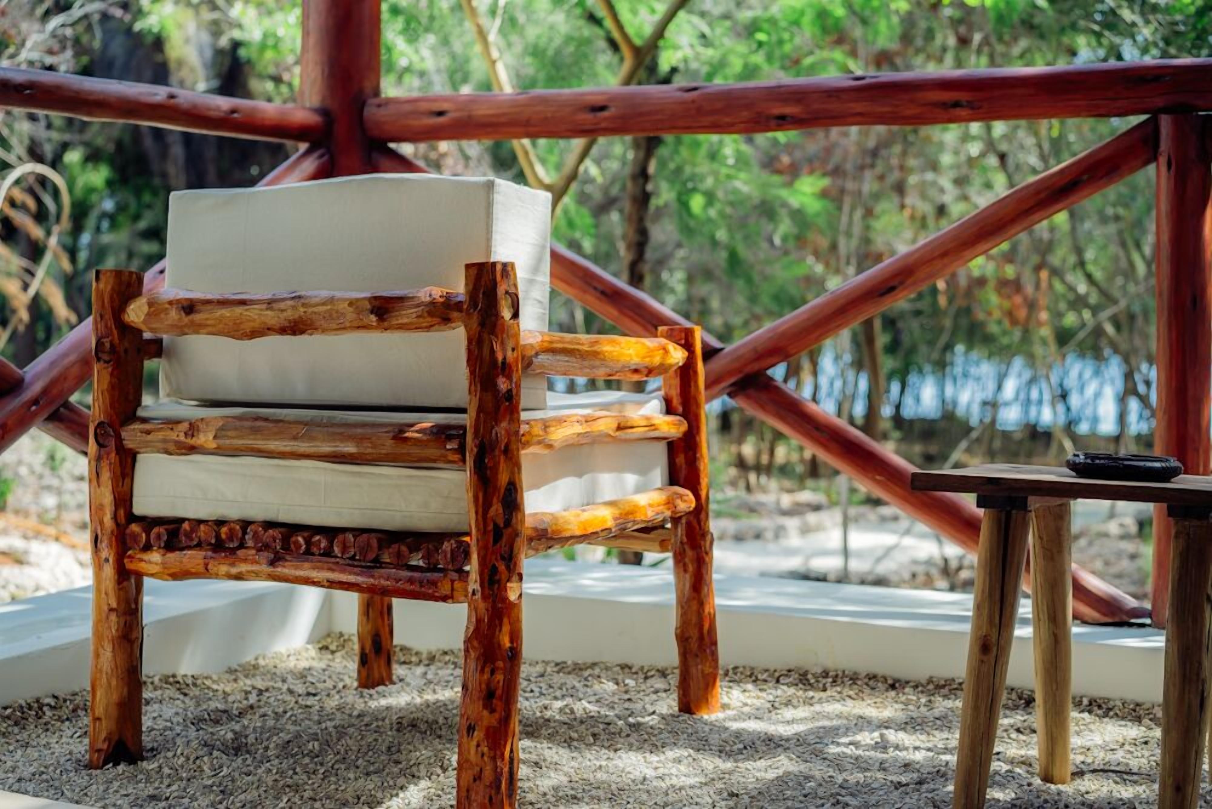 A rustic wooden chair with a cushioned seat, positioned on a gravel surface with a wooden table nearby.