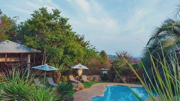 A swimming pool with a stone edge, surrounded by tropical plants and a wooden deck. - Cussonia Country Home (Tierpoort)
