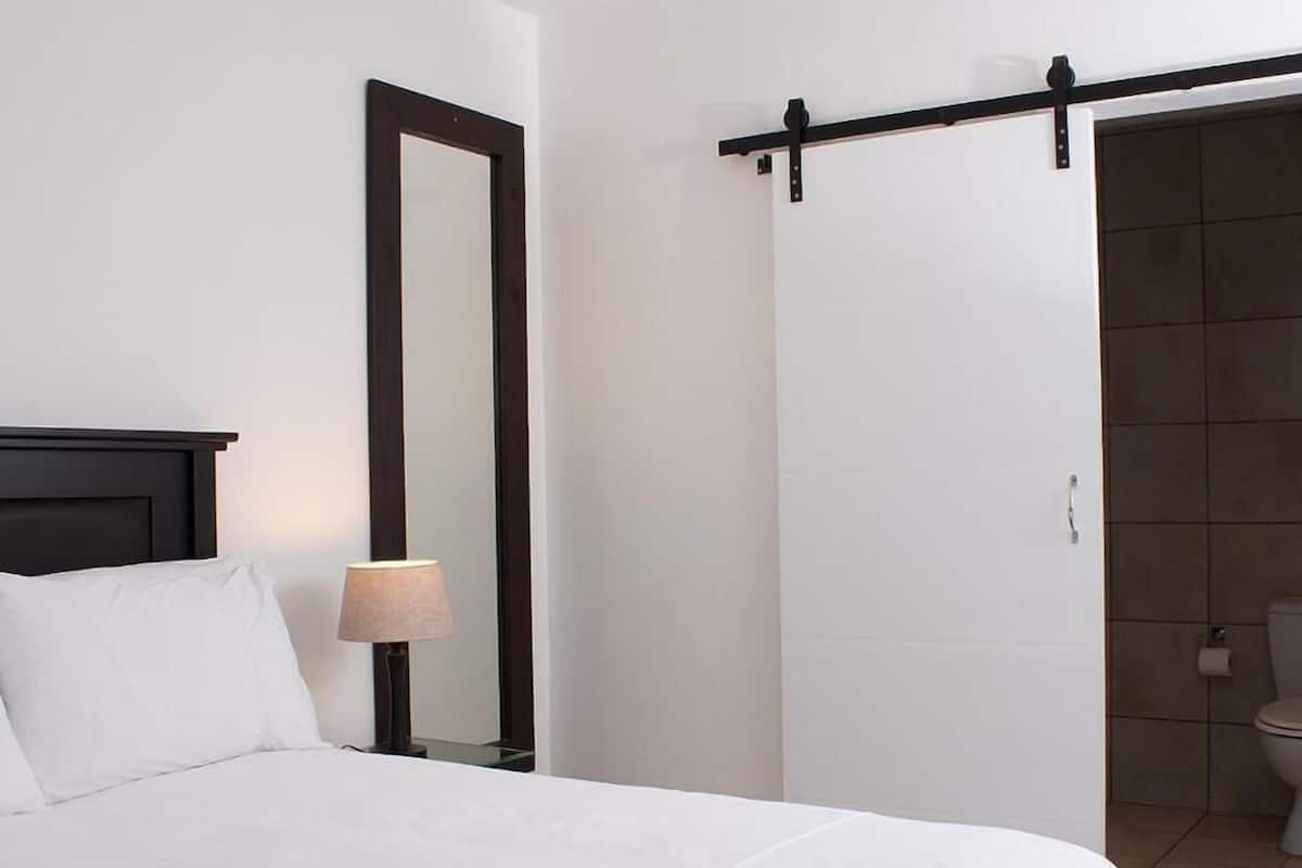 A hotel room with a bed, a nightstand, a lamp, a mirror, and a sliding door leading to a bathroom.