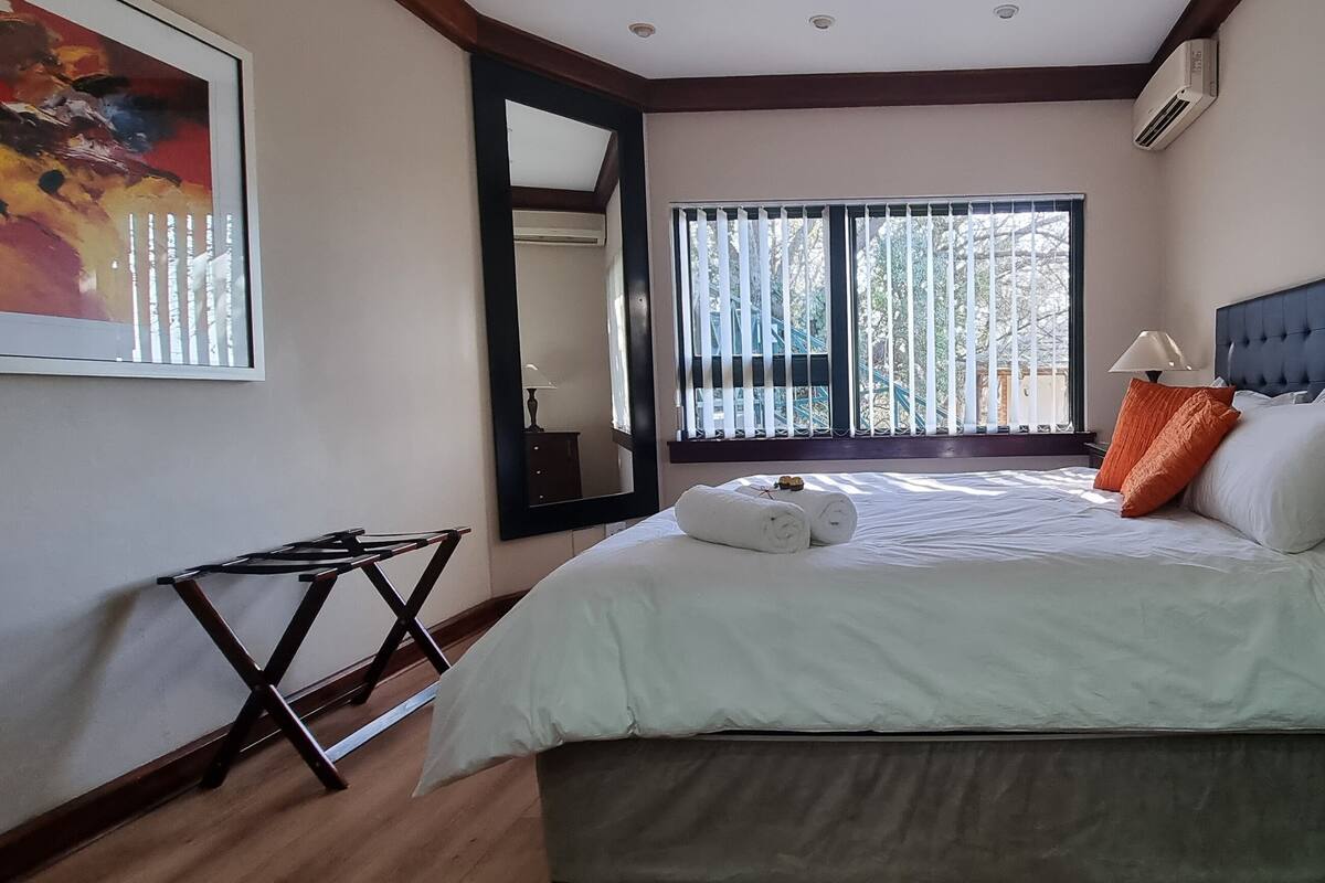 A bedroom with a bed, a nightstand, a lamp, a window with blinds, and a painting on the wall.