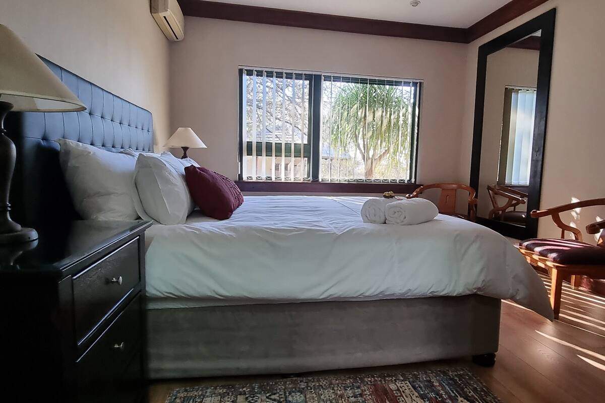 A bedroom with a bed, bedside lamps, a chair, a window with bars, and a painting on the wall.