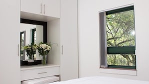 A bedroom with a bed, a dresser with a mirror, a vase with flowers, and a window with a view of trees. - Cussonia Country Home (Tierpoort)