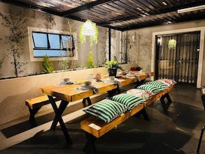 Un espace repas avec une longue table en bois, des bancs, des plantes en pot et des luminaires suspendus.