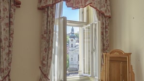 A room with a wooden bed, a blue armchair, a small round table, and a view of a building through an open window with floral curtains. - Hotel Heluan (Karlovy Vary)