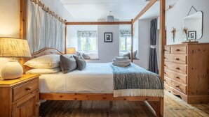 Una cama con dosel de madera, una mesita de noche con lámpara, un aparador con cajones y una ventana con cortinas.