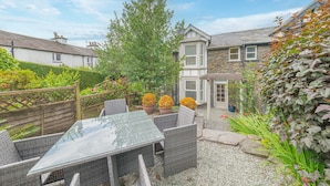 A patio with wicker furniture and a glass table, surrounded by a garden with potted plants and a wooden fence. - Pottery Gate - sleeps 4 in modern dog friendly cottage with parking and new bathroom (Bowness-on-Windermere)