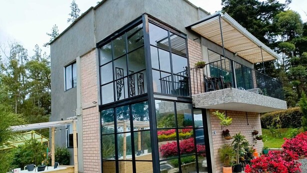 A modern two-story house with large glass windows, a balcony, and a garden with red flowers. - Beautiful villa overlooking the forest + jacuzzi (Guarne)