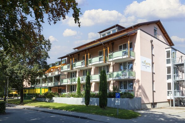 Un hôtel à plusieurs étages, doté de balcons, d’un panneau indiquant l’emplacement du parking et d’un jardin bien entretenu.