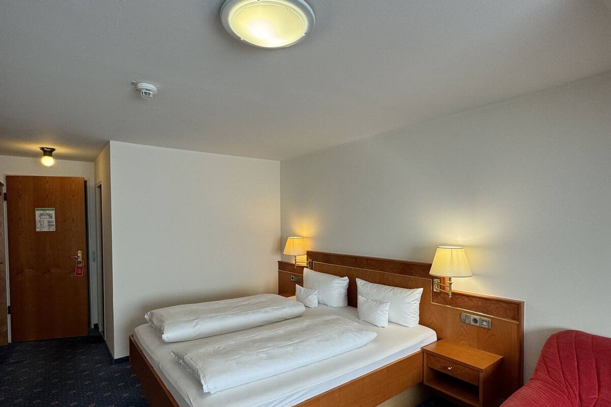 Une chambre d’hôtel avec un grand lit, deux tables de chevet avec des lampes, un fauteuil rouge et une porte.