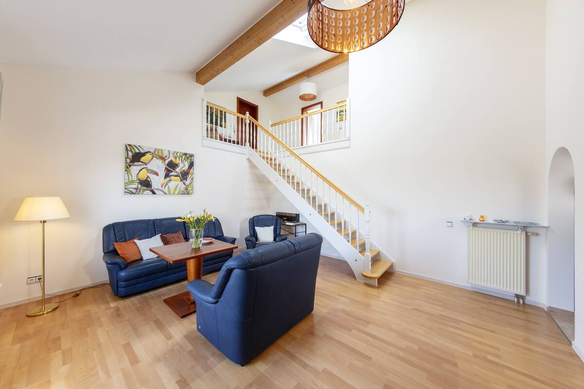 Un vaste espace de vie comprenant un canapé bleu, une table basse en bois et un lampadaire. Un escalier avec une rampe blanche mène à l’étage supérieur.