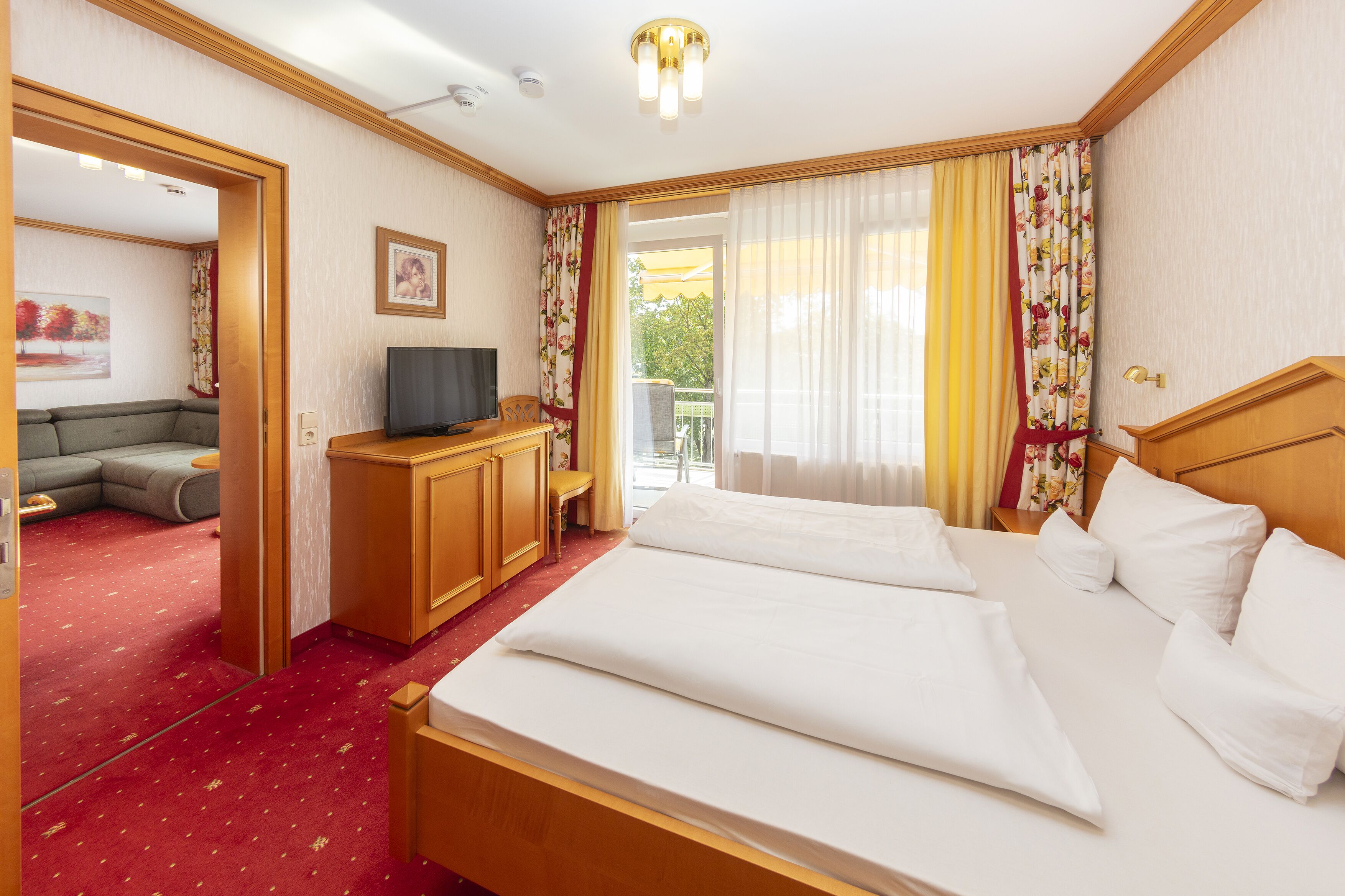 A hotel room with a bed, a TV, a desk, and a balcony with curtains.