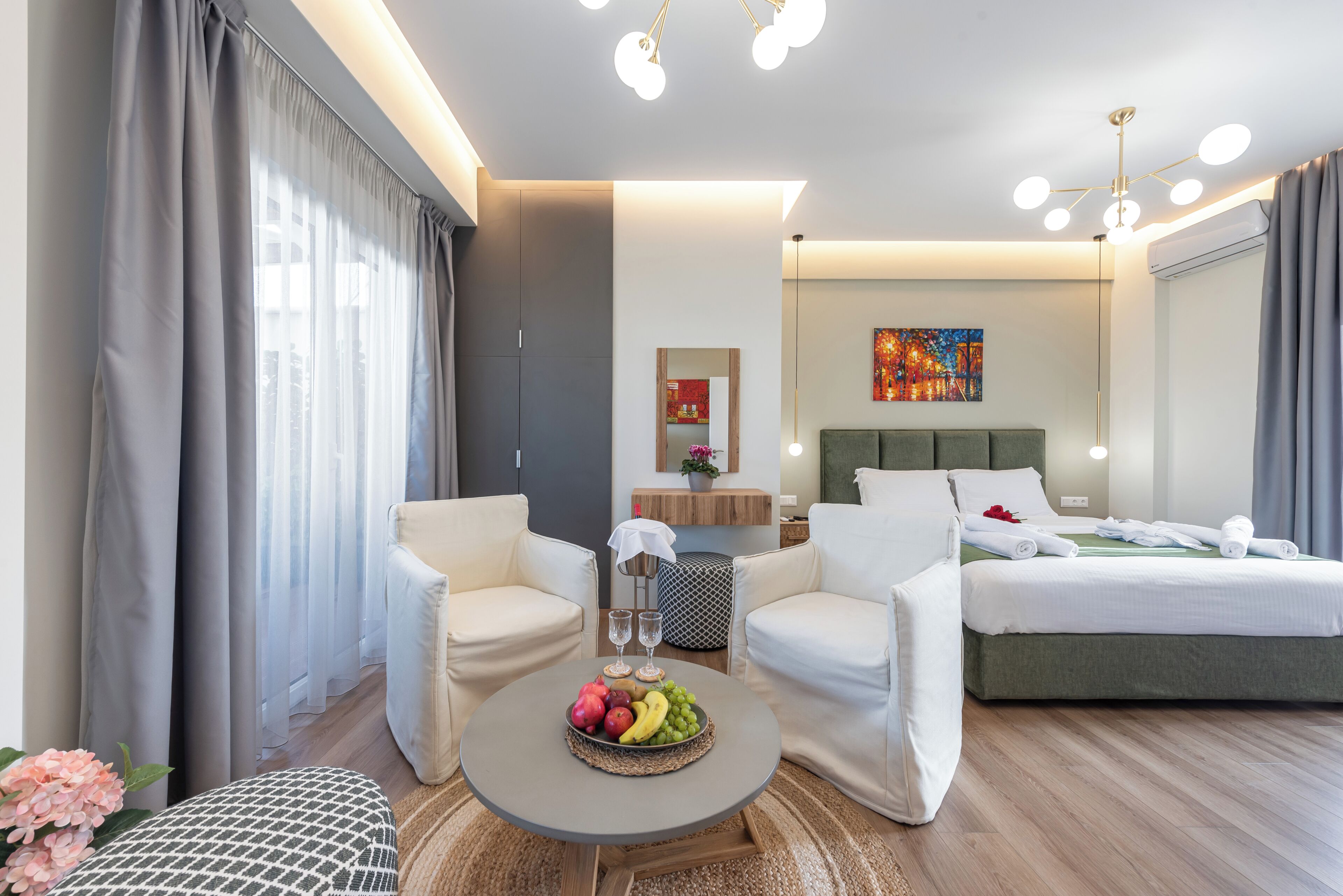 Una habitación de hotel moderna con una cama, dos sillones blancos, una mesa de centro con fruta, una mesita con un florero y un cuadro en la pared.