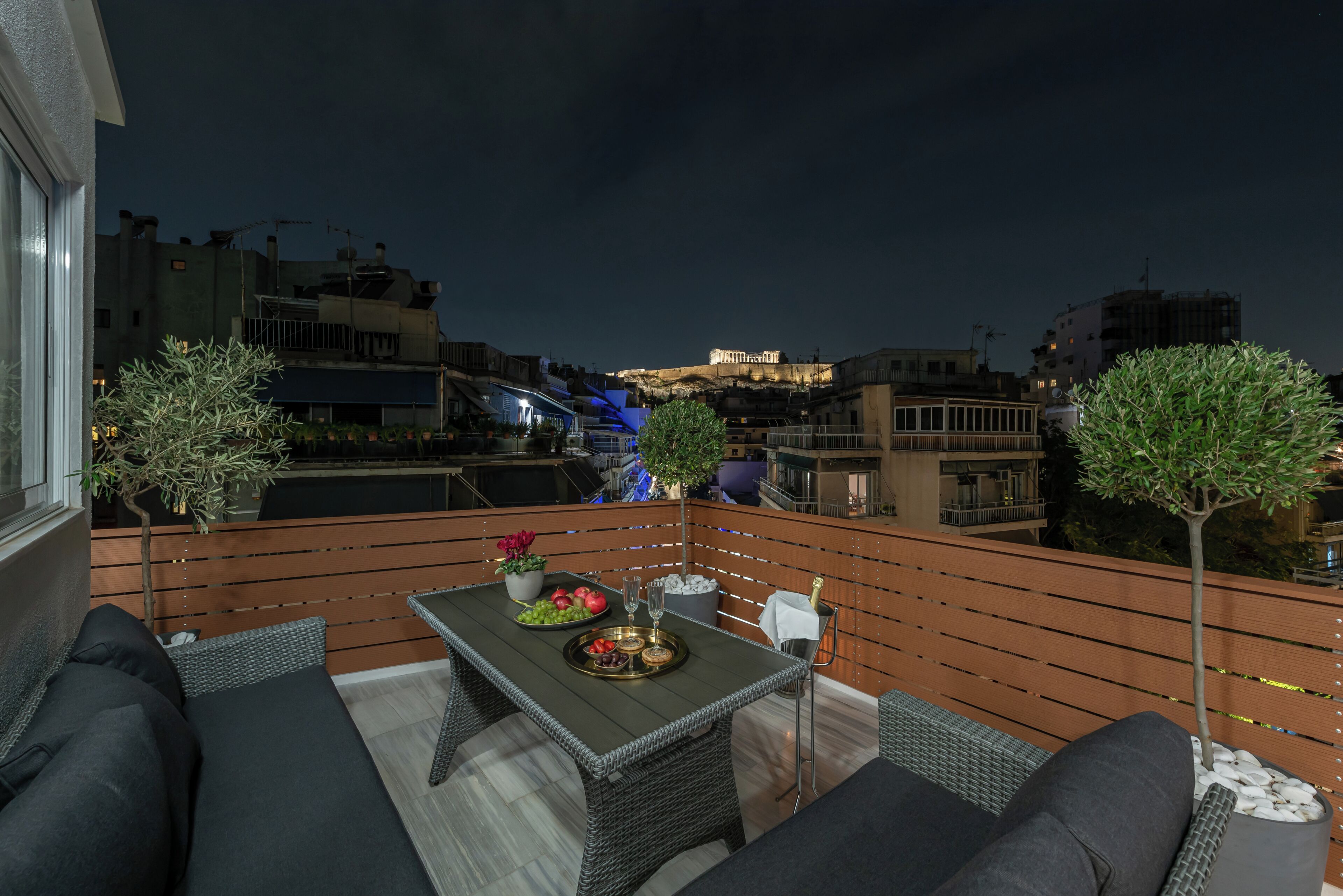 A balcony with wicker furniture, a table with a fruit tray, and a view of a cityscape at night.