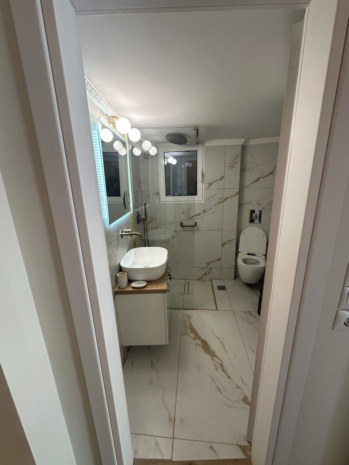 A bathroom with a white sink, mirror, and toilet, marble walls, and a shower area.