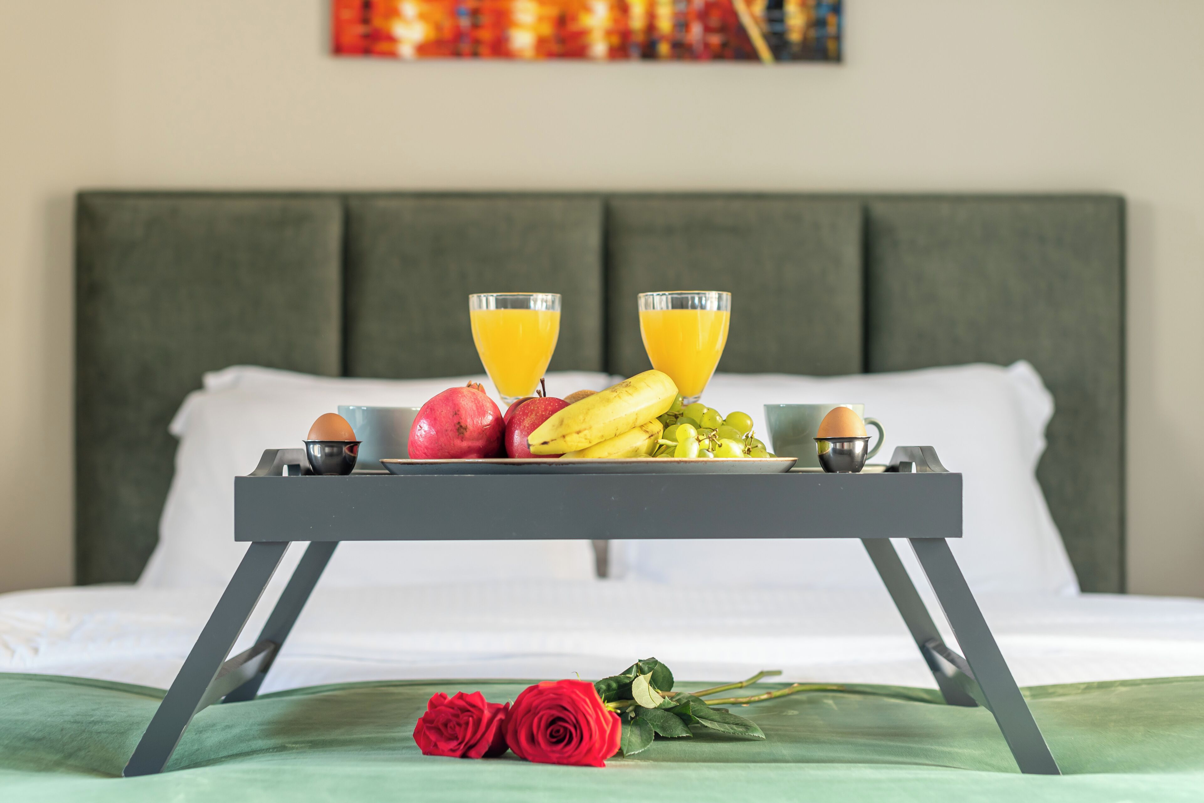 A bed with a tray of breakfast items, including fruits and juice, and two red roses on the bed.