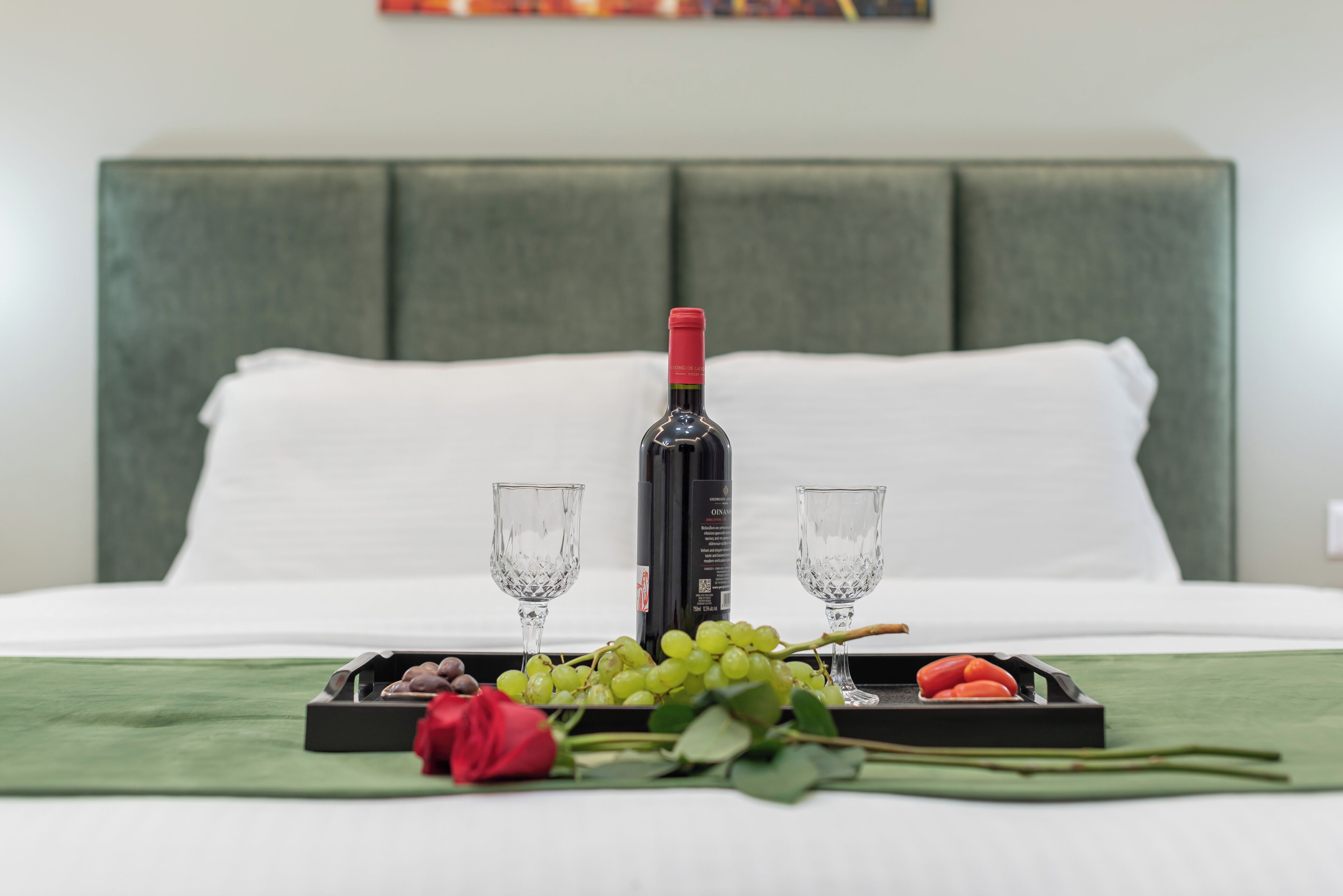 A neatly arranged breakfast tray on a bed with a bottle of wine, two glasses, grapes, and a rose.