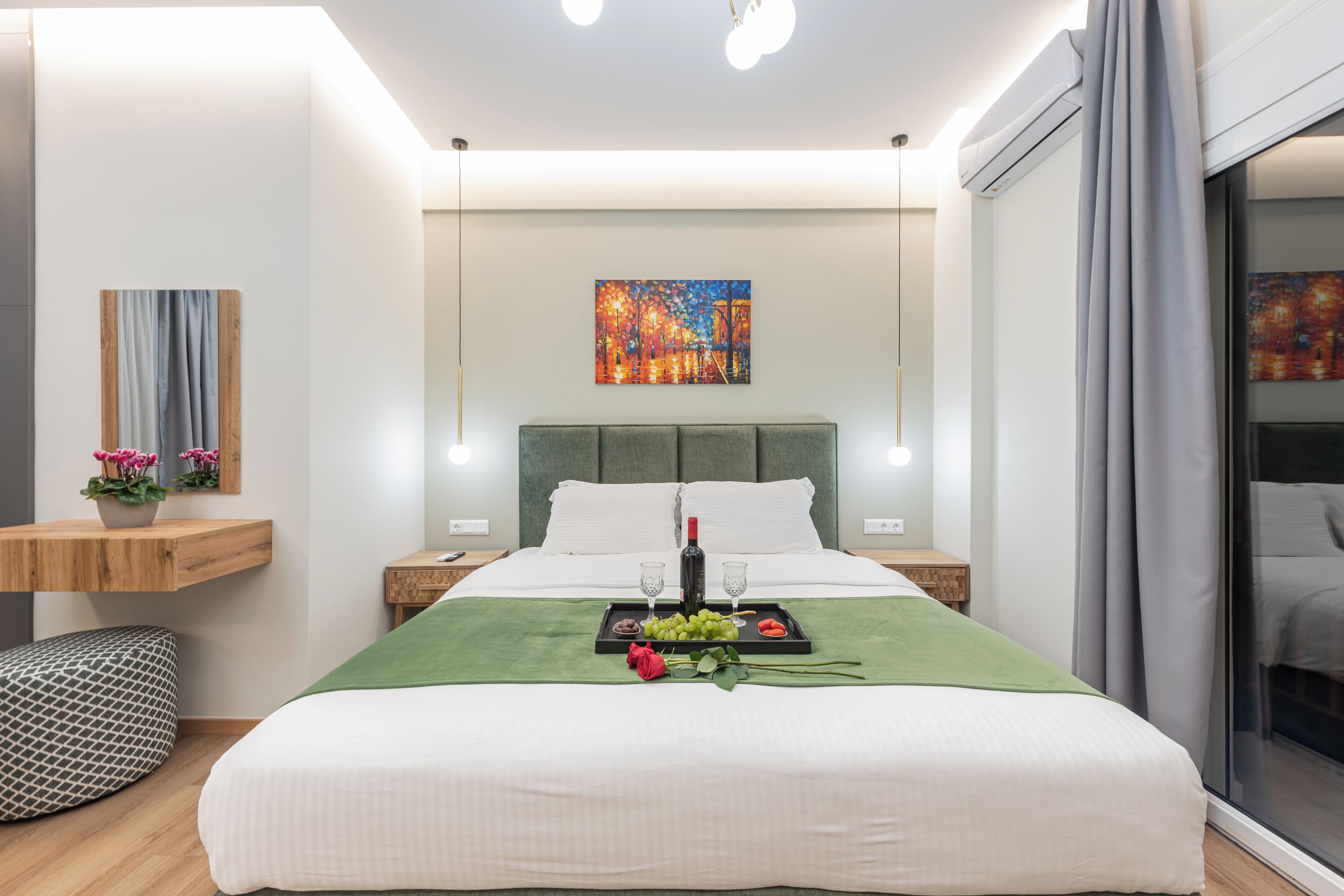 A modern hotel room with a large bed, bedside tables, a green throw blanket, a tray with a bottle, glasses, and fruit.