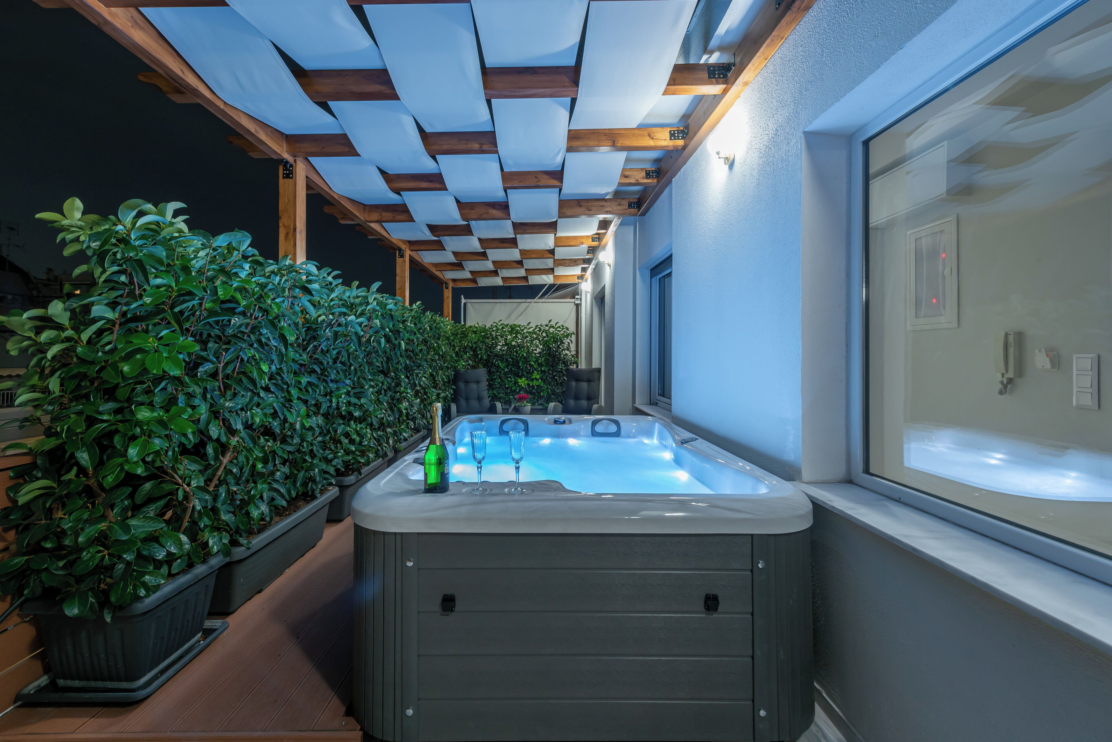 A hot tub with a wooden pergola overhead, potted plants, and a window with a view.