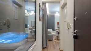 En hall med ett glasinkapslat bubbelbad, ett trägolv, en mörk dörr och utsikt över ett vardagsrum med en vit soffa och ett runt bord.