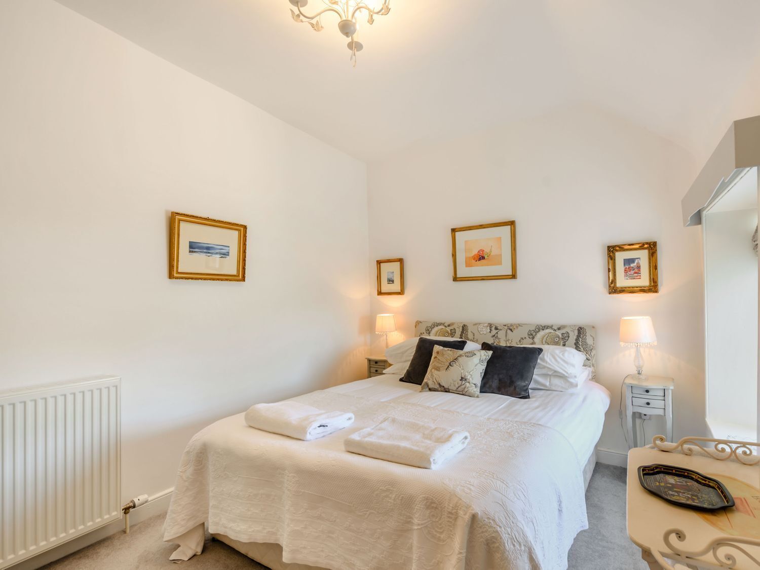 Een net opgemaakt bed met witte lakens en twee handdoeken, een nachtkastje met een lamp, en drie ingelijste foto’s aan de muur.