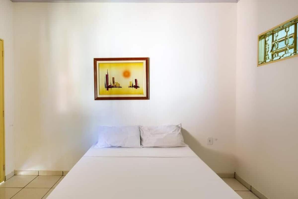 Ein Zimmer mit einem Bett, einem gerahmten Bild an der Wand und einem Fenster mit grünem Rahmen.