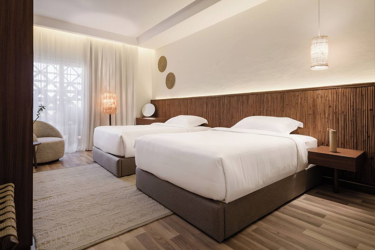 A modern hotel room with two beds, a wooden headboard, a bedside table with a lamp, and a large window with sheer curtains.