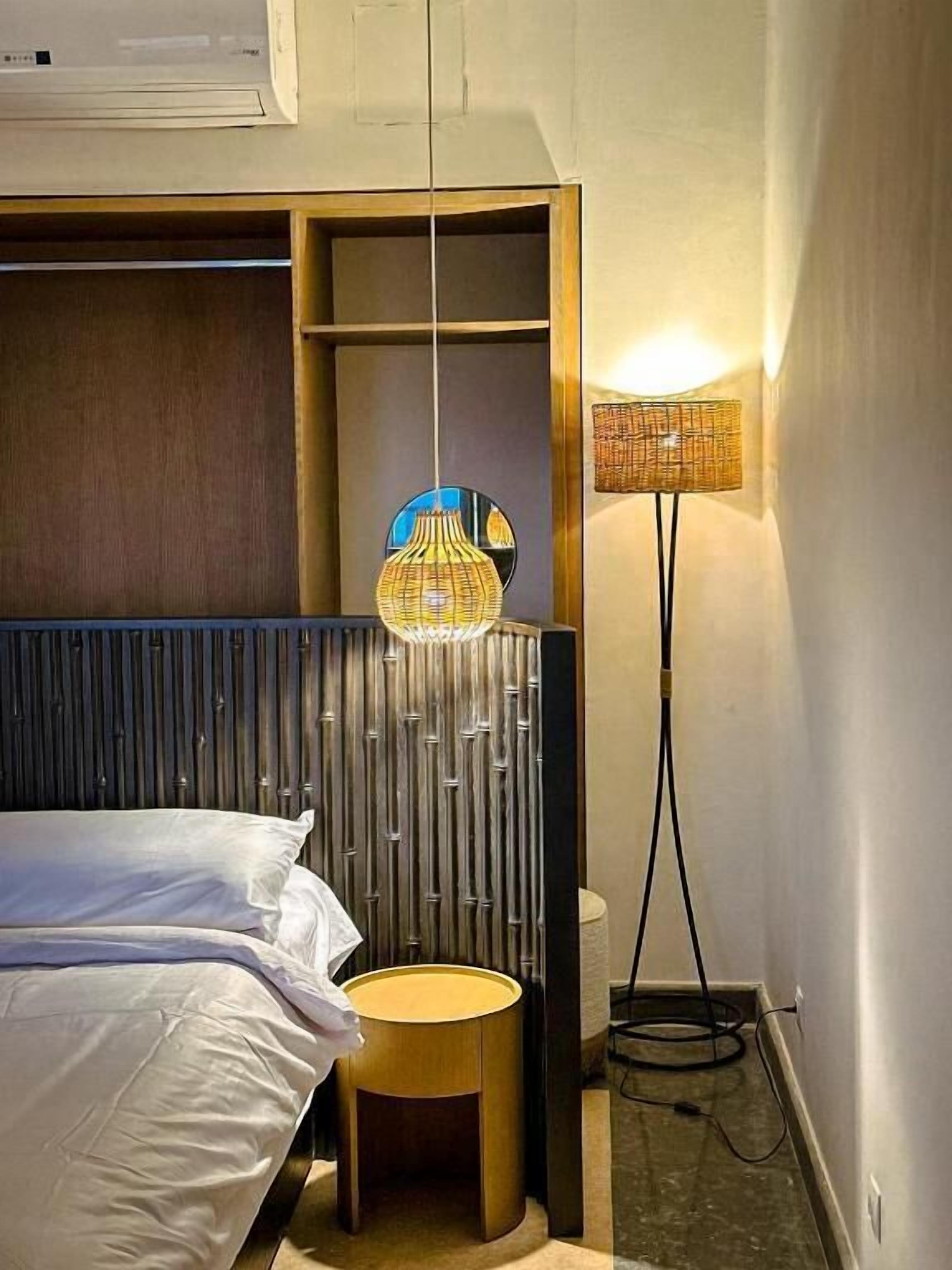 A hotel room with a bed, a round wooden side table, a wall-mounted air conditioner, and a floor lamp.