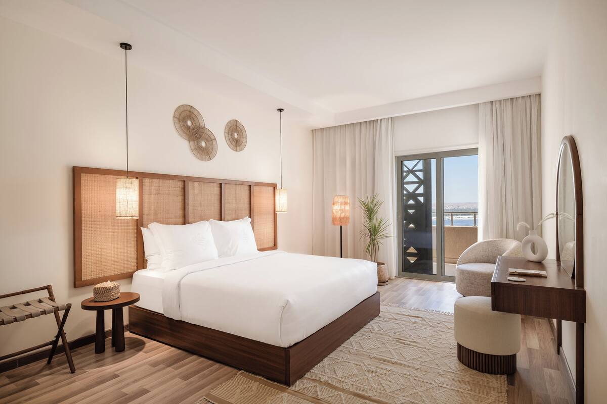 A hotel room with a large bed, a wooden headboard, two wall-mounted lamps, a small round table with a basket, a chair, a desk with a mirror, and a view of the outdoors through a large window.