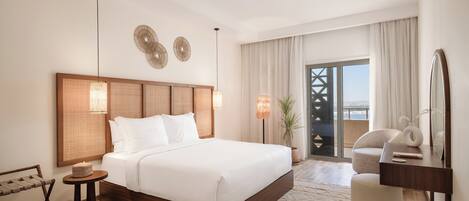A hotel room with a large bed, a wooden headboard, two wall-mounted lamps, a small round table with a basket, a chair, a desk with a mirror, and a view of the outdoors through a large window.