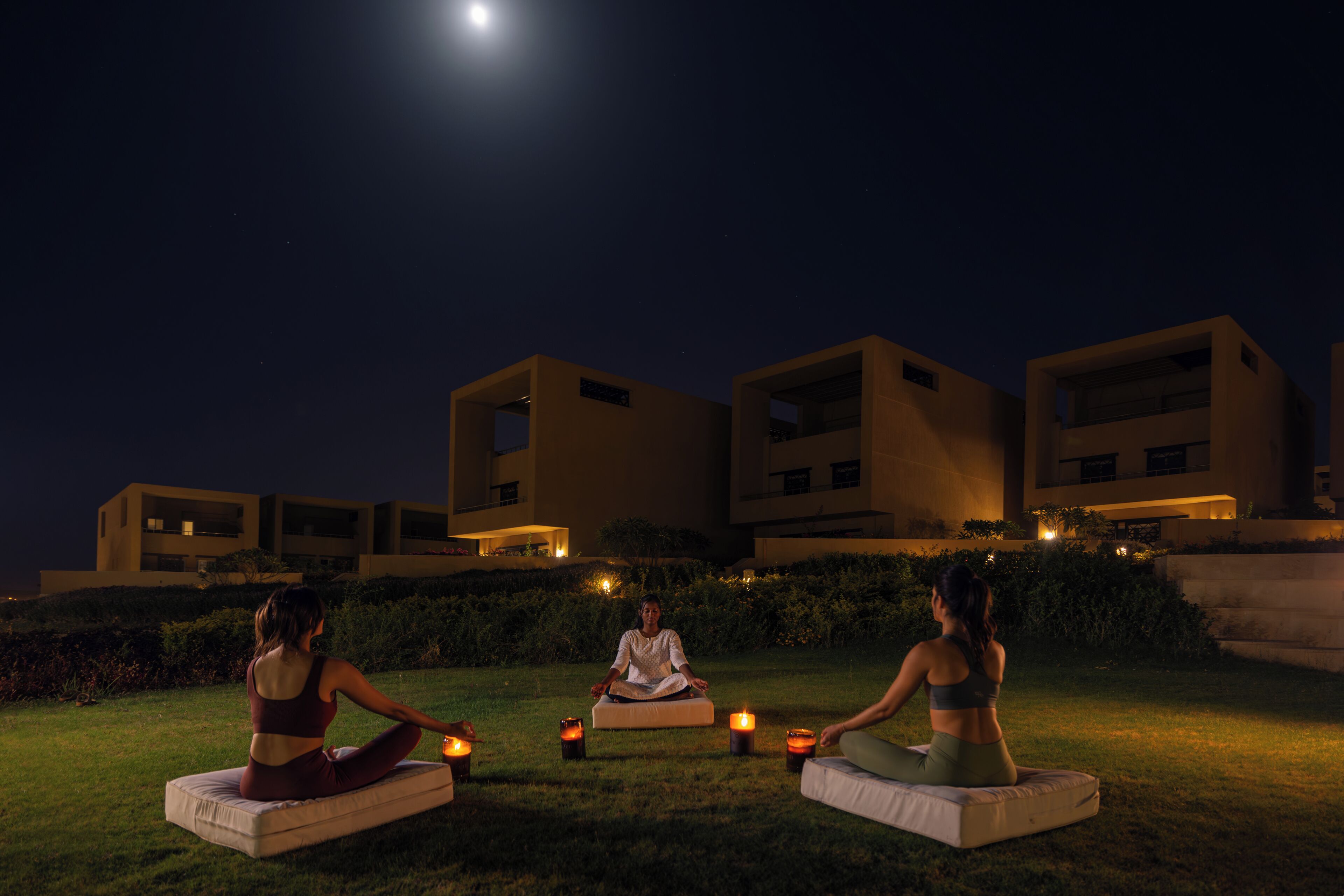 Three people meditating on grass with candles, under a night sky with a visible moon, in front of modern buildings.