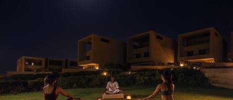 Three people meditating on grass with candles, under a night sky with a visible moon, in front of modern buildings.