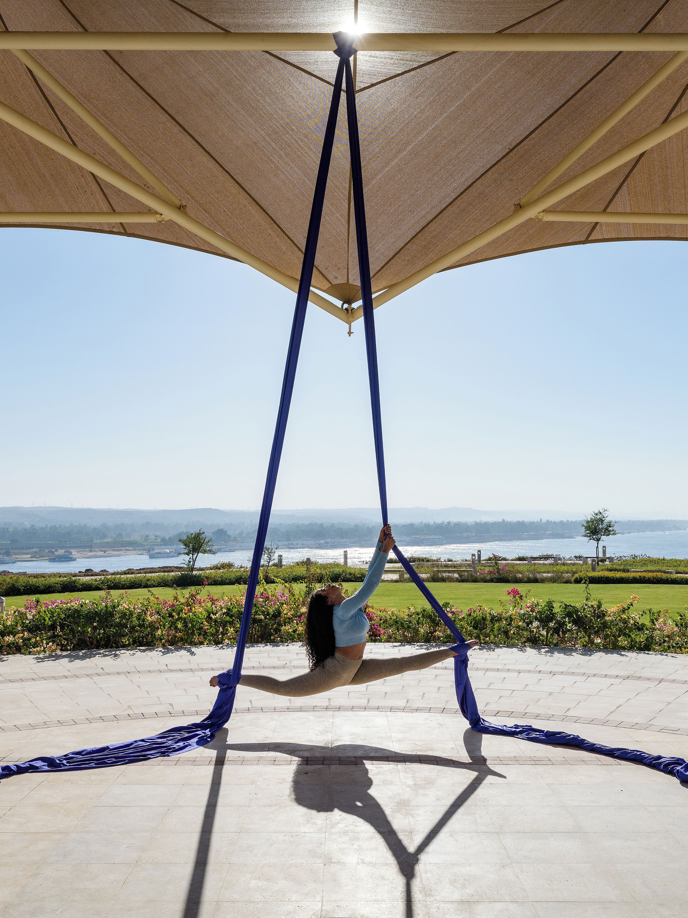 A person performing aerial yoga under a canopy with a scenic view of a body of water and greenery in the background.
