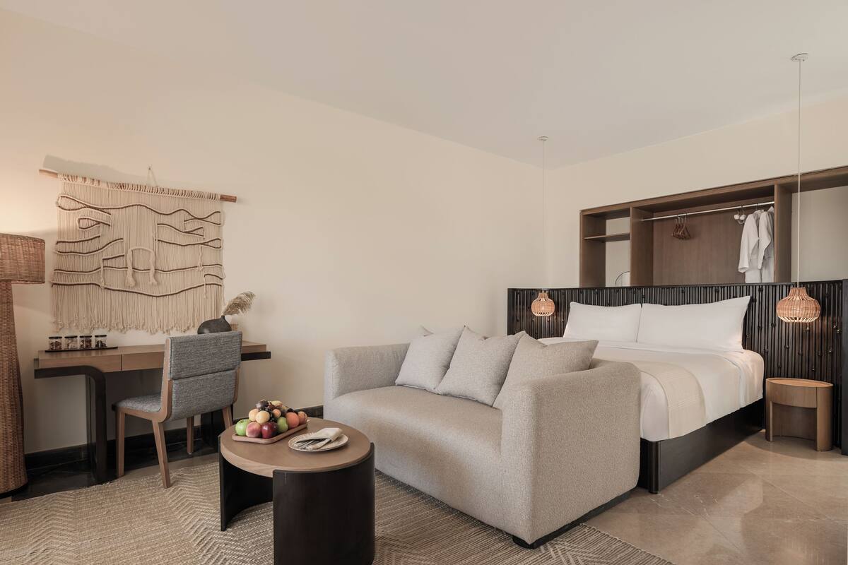 A modern hotel room with a grey sofa, a desk with a chair, a round coffee table, a bed with a wooden headboard, and a large wall-mounted artwork.