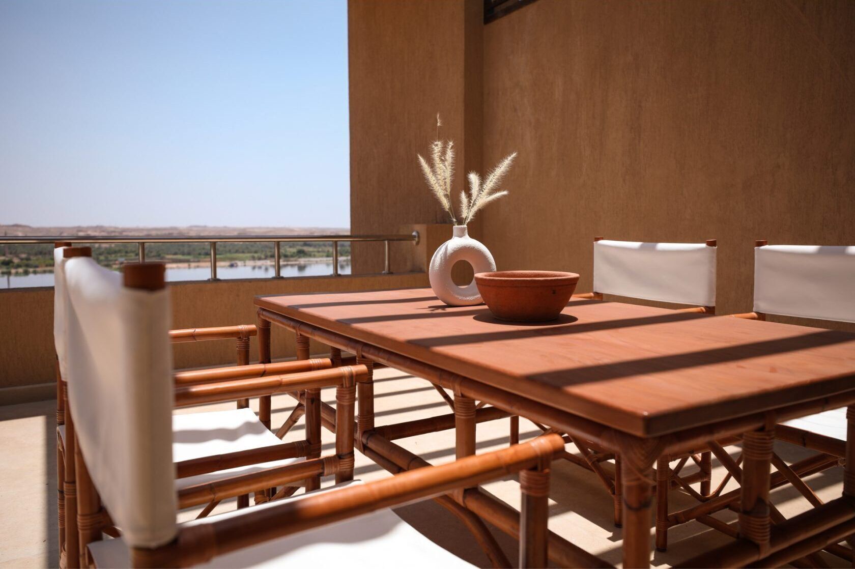 A wooden dining table with chairs, a vase with pampas grass, and a terracotta bowl on a balcony with a view of a body of water and hills.