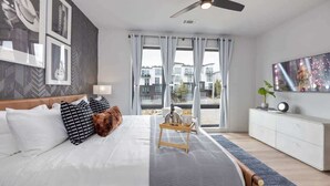 A modern bedroom with a bed, a ceiling fan, a TV, and a view of buildings outside. - Cowboy Condo- Odyssey - Walk to Bars & Restaurants (Nashville)