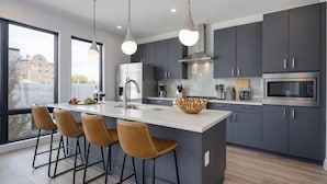 A modern kitchen with a central island, bar stools, pendant lighting, and a view through large windows. - Cowboy Condo- Odyssey - Walk to Bars & Restaurants (Nashville)