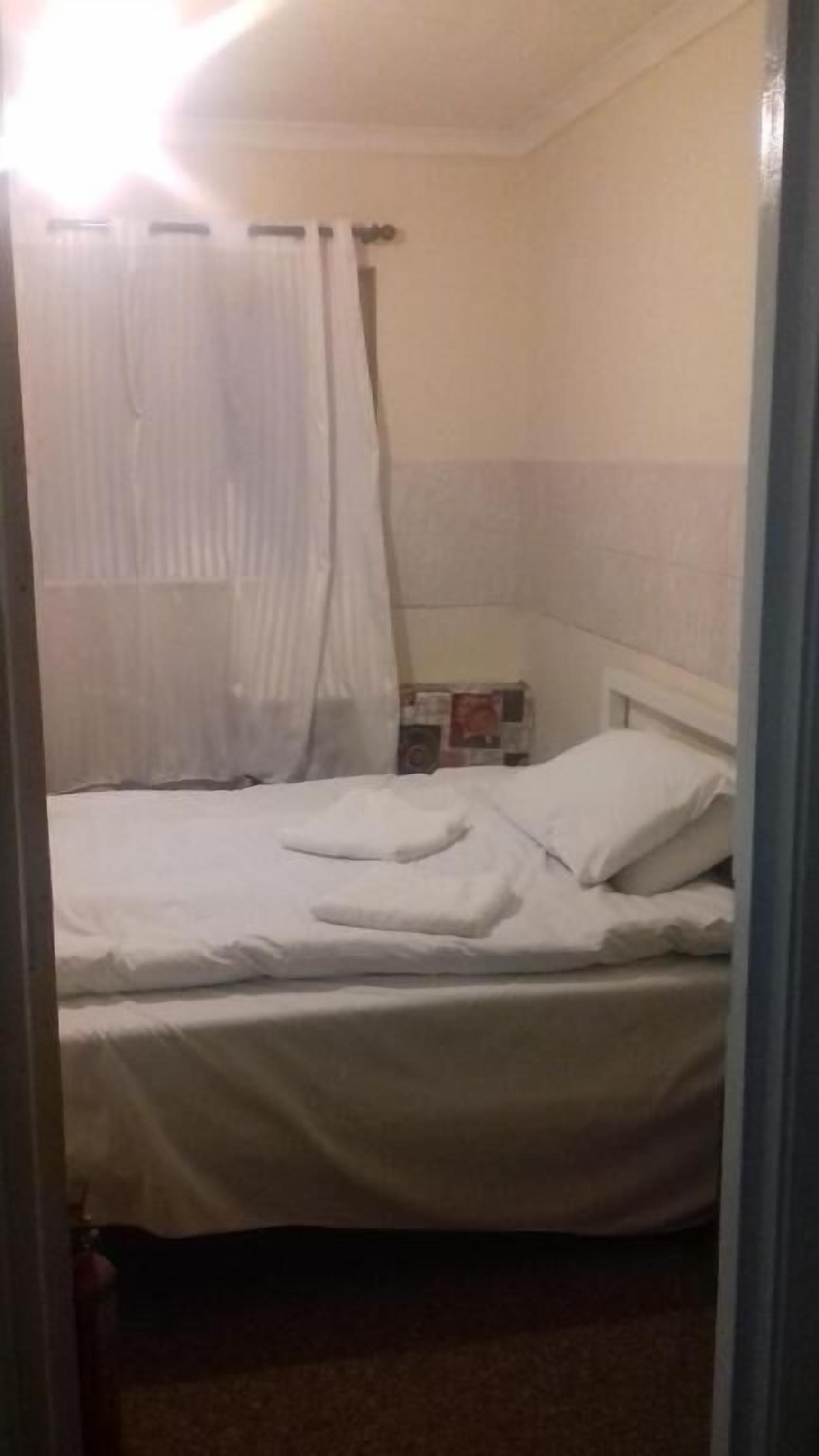 A single bed with white bedding in a room with a plain wall and a curtain.
